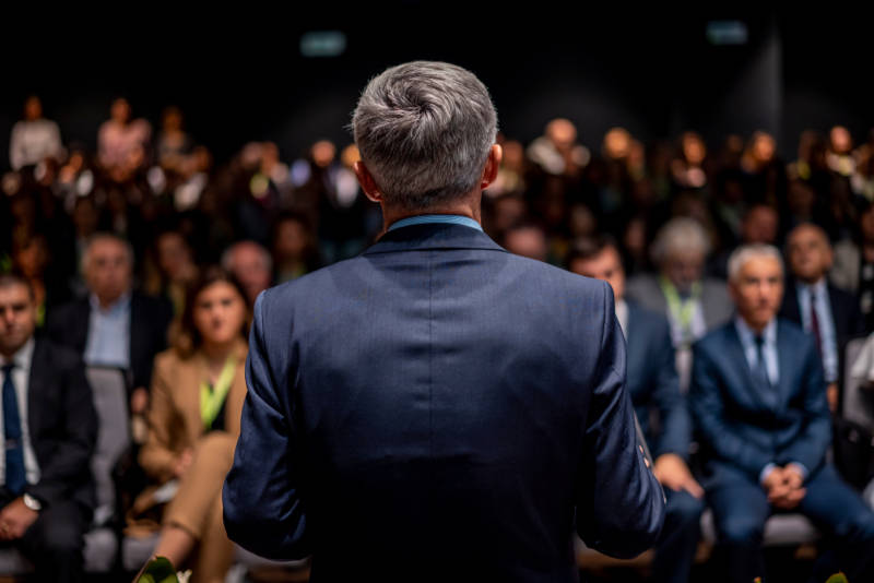 A speaker viewed from the back giving a lecture to a large audience