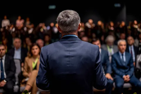 A speaker viewed from the back giving a lecture to a large audience